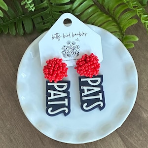 May include: A pair of earrings with red beaded tops and navy blue pendants that read "PATS" in white block letters. The earrings are displayed on a white plate with a tag that reads "betty bird baubles".