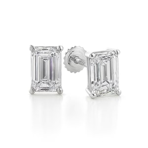 May include: A pair of sparkling emerald-cut diamond stud earrings. Each earring features a rectangular, clear diamond set in a silver or white gold setting. The earrings are displayed on a white background.