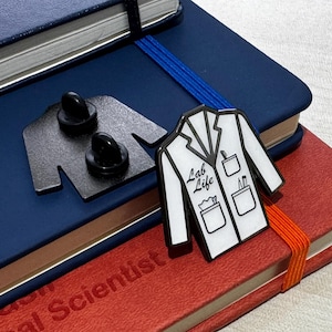 Könnte beinhalten: Eine weiße Emaille-Anstecknadel mit einem Laborkittel-Design. Der Laborkittel hat Taschen mit Stiften und einem Reagenzglas. Der Text "Lab Life" ist auf dem Kittel geschrieben. Die Anstecknadel befindet sich auf einem Stapel Notizbücher.