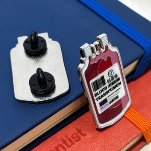 May include: A silver enamel pin shaped like a blood bag with a red and white label that reads "O NEG BLOOD TRANSFUSION".