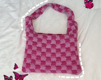 May include: A handmade crochet tote bag in a checkerboard pattern of pink and light purple. The bag has a single handle and is displayed on a white surface. Pink butterflies are scattered around the bag.