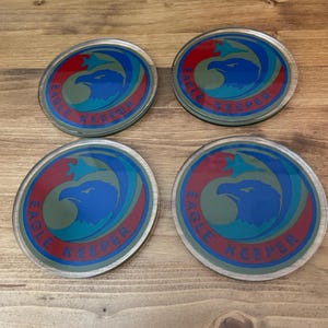 May include: Set of four round coasters featuring a green, blue, and red eagle design. The coasters display the text "Eagle Keeper" in a blue circle around the eagle.