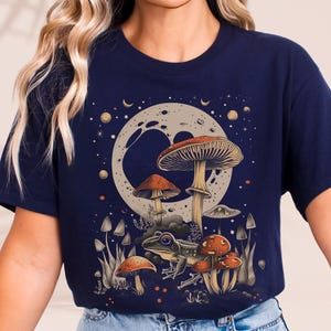 Könnte beinhalten: Marineblaues T-Shirt mit einer fantasievollen Illustration von Pilzen, einem Frosch und einem Mond. Das Design umfasst Pilze in verschiedenen Größen und Farben, einen Frosch mit Brille und einen Mond mit Sternen. Das Shirt ist bequem und lässig.