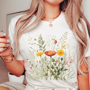 Könnte beinhalten: Weißes T-Shirt mit einem farbenfrohen Blumenmuster. Das Design zeigt eine Auswahl an Wildblumen in Gelb, Orange und Rosa, vor grünem Laub. Das T-Shirt hat einen klassischen Rundhalsausschnitt.