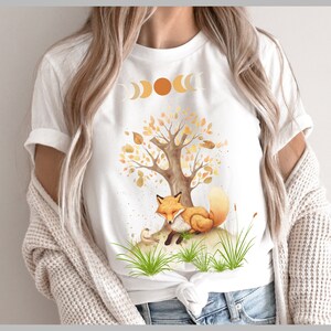 Könnte beinhalten: Weißes T-Shirt mit einem Aufdruck eines schlafenden Fuchses unter einem Baum mit orangefarbenen und gelben Blättern. Der Aufdruck enthält auch eine Mondsichel und eine Sonne.