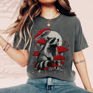 Könnte beinhalten: Dunkelgraues T-Shirt mit einem Waschbären, umgeben von roten Pilzen mit weißen Punkten, vor einem Vollmond und Sternen. Der Waschbär blickt nach oben. Das T-Shirt hat einen Vintage-Look.