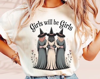 Op de afbeelding: Crèmekleurig T-shirt met de tekst "Girls will be Girls" boven een illustratie van drie figuren in heksenhoeden en jurken. De figuren omarmen elkaar, met bloemaccenten.