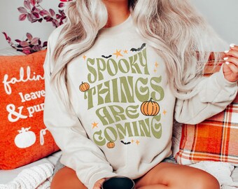May include: Beige sweatshirt with the text "Spooky Things Are Coming" in green, orange, and white. The design includes pumpkins, bats, and stars. The sweatshirt is part of a fall or Halloween-themed collection.