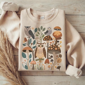 Può includere: Felpa beige con una scena boschiva stravagante con gufi, funghi e fogliame nei toni del marrone, verde e arancione. Il disegno è centrato sulla parte anteriore della felpa, con maniche arrotolate.