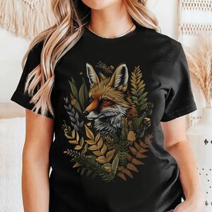 Könnte beinhalten: Schwarzes T-Shirt mit einer detaillierten Illustration eines Fuchses, der von Blättern umgeben ist. Der Fuchs hat orangefarbenes, braunes und weißes Fell, mit grünen, goldenen und grauen Blättern und Pflanzen. Das Design ist auf dem Shirt zentriert.