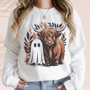 Könnte beinhalten: Weißes Sweatshirt mit einem braunen Highland-Kuh- und einem weißen Geister-Design. Die Kuh hat große Hörner und ist von Blättern umgeben. Der Geist hat zwei schwarze Augenlöcher. Das Design ist auf der Vorderseite des Sweatshirts zentriert.