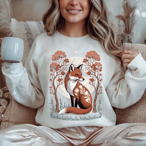 Könnte beinhalten: Cremefarbenes Sweatshirt mit einer detaillierten Illustration eines Fuchses mit floralen Akzenten, vor dem Hintergrund von Bäumen und einem Vollmond. Der Fuchs ist in Braun-, Weiß- und Schwarztönen dargestellt.