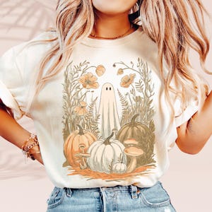 Könnte beinhalten: Cremefarbenes T-Shirt mit einem skurrilen Halloween-Design. Die Grafik zeigt einen Geist, Kürbisse, Pilze und florale Elemente in Orange-, Braun- und Weißtönen. Das Design hat eine Vintage-Ästhetik mit leicht verblassten Farben.