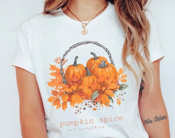 May include: White t-shirt featuring a watercolor illustration of pumpkins in a woven basket, surrounded by orange autumn leaves. The text "pumpkin spice and everything nice" is printed below the image, evoking a fall theme.
