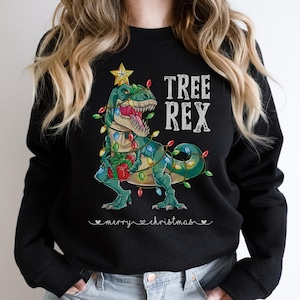 Könnte beinhalten: Schwarzes Sweatshirt mit einem Cartoon-Dinosaurier, der als Weihnachtsbaum geschmückt ist, mit bunten Lichtern umwickelt ist und ein Geschenk hält. Der Text "TREE REX" steht über dem Dinosaurier, darunter "merry christmas".