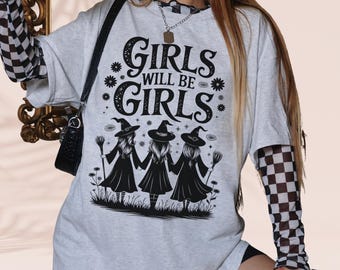Op de afbeelding: Lichtgrijs T-shirt met de tekst "GIRLS WILL BE GIRLS" en een afbeelding van drie heksenfiguren. Het shirt heeft korte mouwen en is gelaagd over een geruit shirt met lange mouwen.