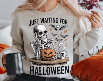 May include: Beige sweatshirt with a Halloween-themed graphic. The design features a skeleton, a carved pumpkin, bats, and the text "JUST WAITING FOR HALLOWEEN". A black mug is held in the left hand.