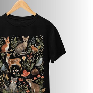 Könnte beinhalten: Schwarzes T-Shirt mit einer verspielten Waldszene. Das Design zeigt verschiedene Waldtiere wie ein Reh, Fuchs, Hase und Vogel, umgeben von floralen und Blattelementen. Die Illustration ist in sanften, natürlichen Farben gehalten.