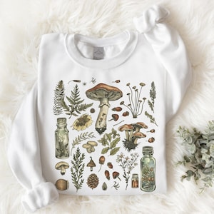 Peut inclure: Sweat-shirt blanc orné d'une illustration botanique détaillée de champignons, de plantes et de feuillages. Le motif comprend un grand champignon à chapeau brun, plusieurs petits champignons, des fougères et d'autres éléments naturels.