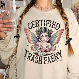 Könnte beinhalten: Beiges Sweatshirt mit einem Grafik einer Fee mit Regenbogenflügeln und dem Text "CERTIFIED TRASH FAERY". Die Fee hat ein Rosen-Tattoo auf dem Arm. Das Sweatshirt ist ein Rundhals-Stil.