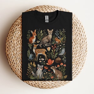 Könnte beinhalten: Schwarzes T-Shirt mit einer Aquarellillustration von Waldtieren und Pflanzen. Das Design umfasst einen Fuchs, ein Reh, ein Kaninchen, ein Eichhörnchen und Vögel, umgeben von Blumen und Blättern. Das T-Shirt ist auf einer gewebten Oberfläche gefaltet.