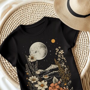 Könnte beinhalten: Schwarzes T-Shirt mit einem Himmelsdesign mit Vollmond, Sternen und floralen Elementen in Weiß, Pfirsich und Beige. Das Shirt hat einen runden Ausschnitt und kurze Ärmel. Ein Strohhut ist im Hintergrund zu sehen.