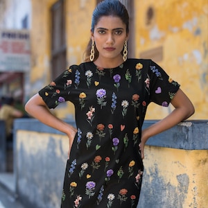 Könnte beinhalten: Ein schwarzes, kurzärmeliges Kleid mit einem Blumendruck, der verschiedene farbige Blumen und Herzen zeigt. Das Kleid hat einen runden Ausschnitt und eine lockere Passform.