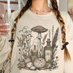 Könnte beinhalten: Beiges Sweatshirt mit einer detaillierten Illustration von Pilzen, Pflanzen und Flaschen. Das Design zeigt einen großen Pilz in der Mitte, umgeben von kleineren Pilzen und botanischen Elementen. Ein Stern befindet sich auf einer der Flaschen.