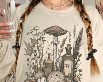 May include: Beige sweatshirt featuring a detailed illustration of various mushrooms, plants, and bottles. The design includes a large mushroom in the center, surrounded by smaller fungi and botanical elements. A star is on one of the bottles.