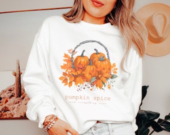 May include: White sweatshirt featuring a watercolor illustration of pumpkins in a woven basket with fall foliage. The text "pumpkin spice and everything nice" is printed below the image. The design incorporates orange, brown, and green colors.