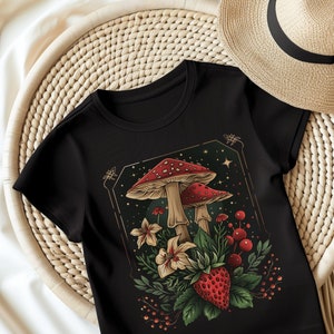 Op de afbeelding: Zwart T-shirt met een grillige illustratie van rood-gekapte paddenstoelen, bloemen en een aardbei in een goudkleurige lijst. Het ontwerp bevat sterren en bessen, wat een op de natuur geïnspireerde esthetiek creëert.