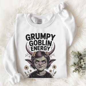 Könnte beinhalten: Weißes Sweatshirt mit dem Text "GRUMPY GOBLIN ENERGY" über einer Cartoon-Goblin-Illustration. Der Goblin hat große Ohren, Hörner und einen Blumenkranz. Gelbe Blumen und Grünzeug befinden sich am unteren Rand des Designs.