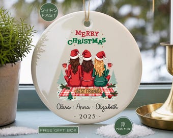 May include: A round white ceramic Christmas ornament featuring an illustration of three people in Santa hats sitting on a red plaid blanket. The text reads Merry Christmas, Best Friends, the names Clara, Anna, Elizabeth, and the year 2025.