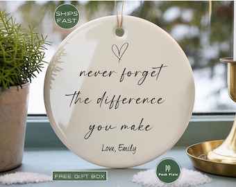 May include: A round, white ceramic ornament featuring a black heart icon and the black printed text: never forget The difference you make Love, Emily. The ornament has a small hole at the top with a thin gold string for hanging purposes.