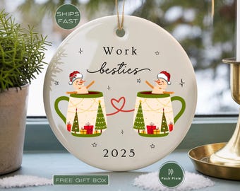 May include: A round white ceramic ornament featuring the text Work besties and 2025. The design shows two snowmen wearing Santa hats, each sitting in a mug decorated with Christmas trees and gifts, connected by a red heart-shaped line.