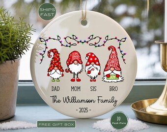 May include: A round white ceramic Christmas ornament featuring four gnomes with red hats, string lights, and the text Dad, Mom, Sis, Bro, The Williamson Family, and 2025. The ornament has a gold hanging string and a smooth, glossy finish.