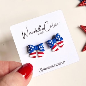 May include: A pair of bow-shaped earrings with a patriotic design, featuring blue bows with white polka dots and red and white striped ribbons. The earrings are displayed on a white card with the text "Wandering Calico Clay". Decorative red, white, and blue star-shaped ornaments are in the background.