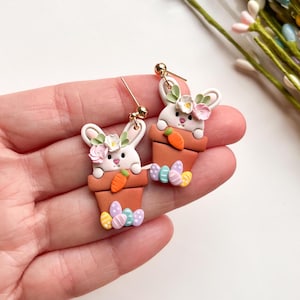 Puede incluir: Un par de pendientes colgantes de oro con un conejo blanco con una corona de flores, asomándose a una maceta marrón con una zanahoria y huevos de Pascua de colores pastel.