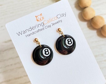 May include: A pair of black and white 8-ball earrings with gold hooks. The earrings are handmade and feature a black clay base with a white 8 painted on it. The earrings are displayed on a white card with the text "WanderingCalicoClay Handcrafted Clay Jewelry" and the Instagram handle "@wanderingcalicoclay".