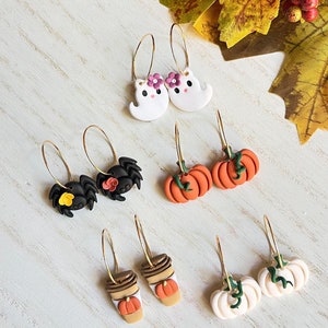 May include: A collection of Halloween-themed earrings. The earrings feature designs such as ghosts with flower crowns, spiders with flowers, orange pumpkins, coffee cups with pumpkins, and white pumpkins. All earrings have gold hoops.