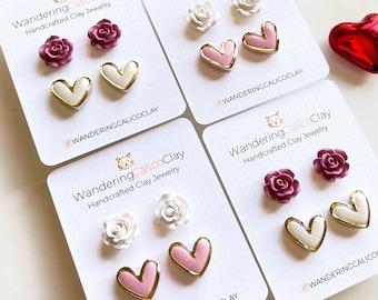 May include: Four sets of handmade clay earrings. Each set includes two earrings. The first set features two pink hearts with gold trim. The second set features two white hearts with gold trim. The third set features two white roses with gold trim. The fourth set features two pink roses with gold trim. Each set is displayed on a white card with the text "WanderingCalicoClay Handcrafted Clay Jewelry" and the Instagram handle "@wanderingcalicoClay".