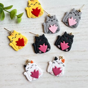 May include: Six pairs of cat-shaped earrings with a heart design. The cats are in various colors, including yellow, gray, black, and white. Each cat is holding a heart in its paws.