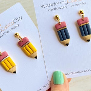 May include: Handcrafted clay jewelry earrings shaped like pencils. The earrings have a yellow body, a pink eraser, and a black tip. The earrings are on a white card with the text "Wandering Calico Clay".