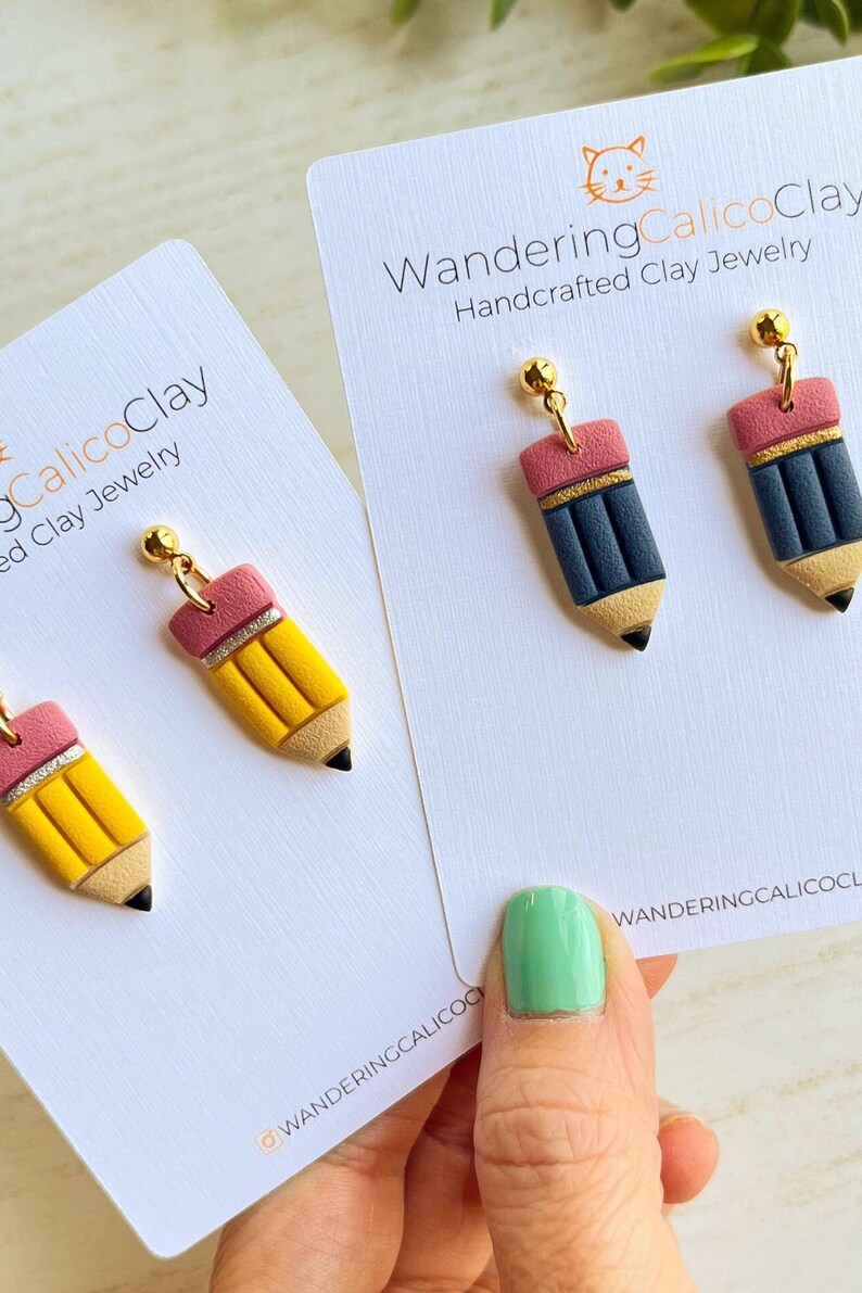 May include: Handcrafted clay jewelry earrings shaped like pencils. The earrings have a yellow body, a pink eraser, and a black tip. The earrings are on a white card with the text "Wandering Calico Clay".