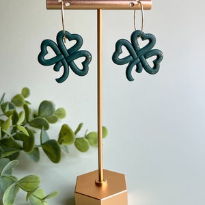 May include: A pair of green shamrock earrings with gold hoops hanging from a gold earring stand.
