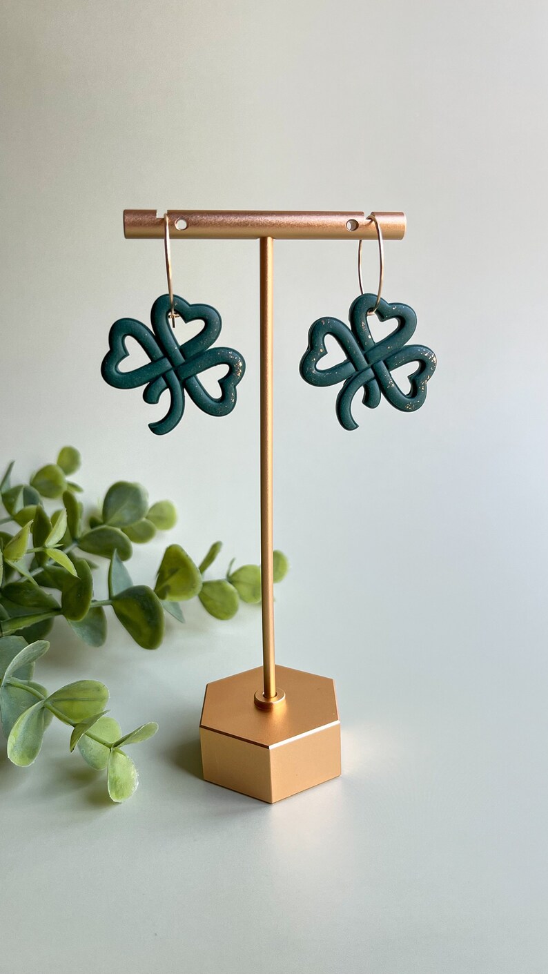 May include: A pair of green shamrock earrings with gold hoops hanging from a gold earring stand.