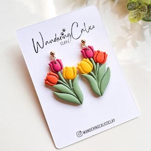 Puede incluir: Un par de pendientes de arcilla polimérica con un grupo de tulipanes coloridos con hojas verdes. Los pendientes están en una tarjeta blanca con el texto "Wandering Calico Clay" y un nombre de usuario de redes sociales.