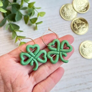 May include: A pair of gold hoop earrings with green shamrock charms. The shamrocks are made of a textured material and have a knotted design.