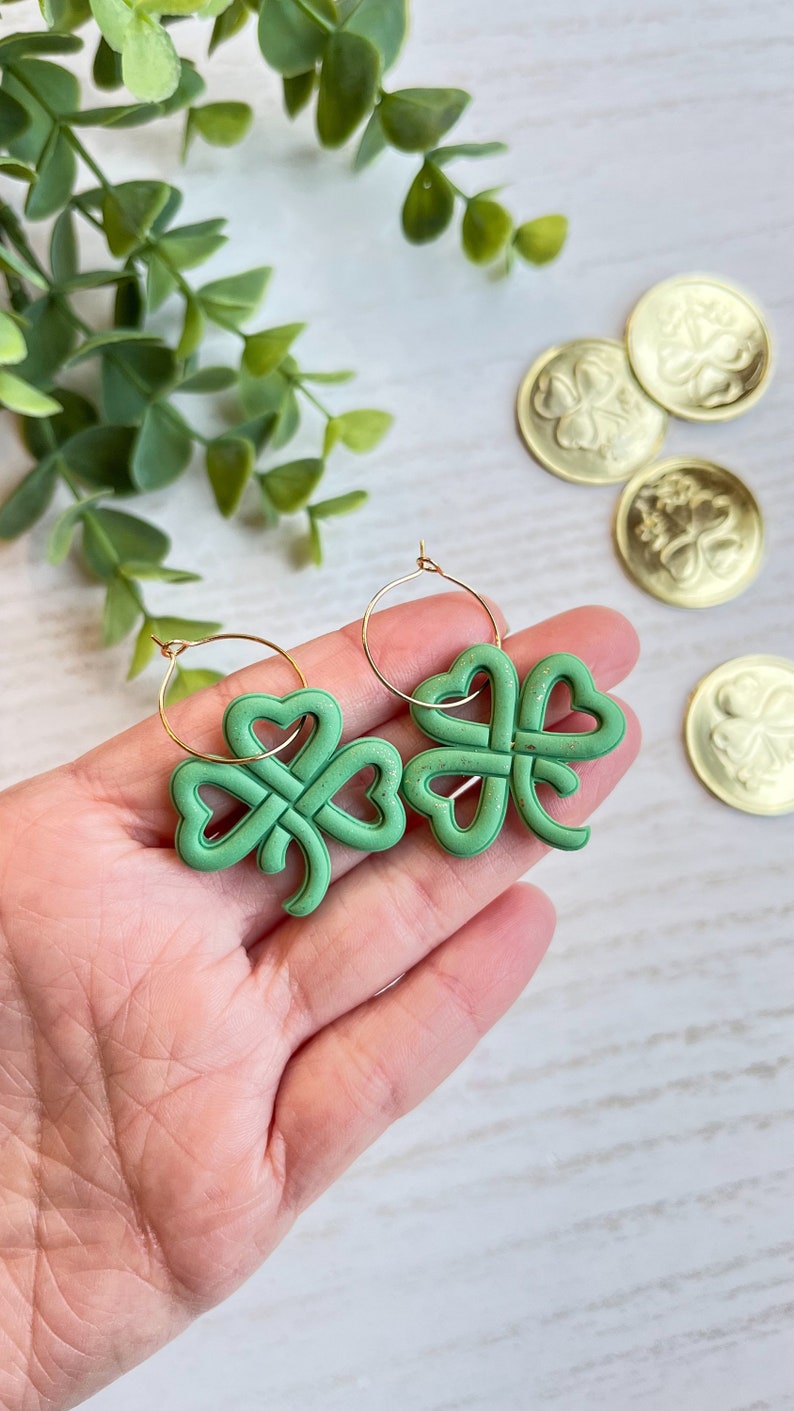 May include: A pair of gold hoop earrings with green shamrock charms. The shamrocks are made of a textured material and have a knotted design.