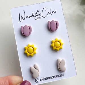 Puede incluir: Un conjunto de seis pendientes de arcilla polim&eacute;rica con tres dise&ntilde;os diferentes: una flor rosa, un sol amarillo y una mariposa blanca. Los pendientes se muestran en una tarjeta blanca con el texto "Wandering Calico Clay" y un nombre de usuario de Instagram.